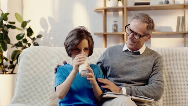 Boy drinking milk near happy middle aged grandfather in eyeglasses with book — стоковое фото