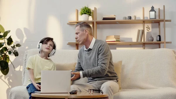 Middle aged grandfather holding book near grandson listening music  in headphones — стоковое фото