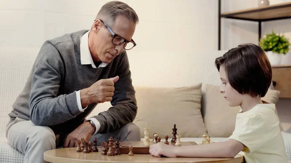 Abuelo en gafas y nieto inteligente jugando ajedrez en la sala de estar - foto de stock