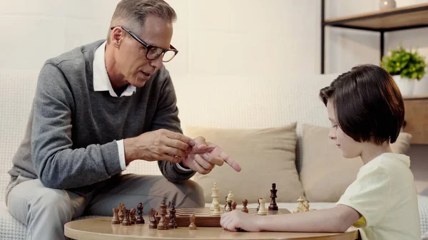 Grand-père dans des lunettes pointant vers la figure tout en jouant aux échecs avec le garçon dans le salon — Photo de stock
