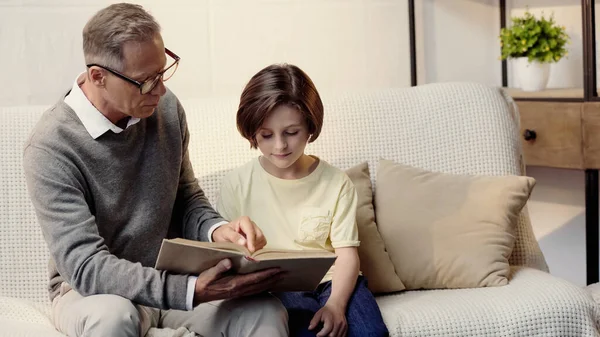 Abuelo de mediana edad en gafas sosteniendo libro cerca de nieto en casa - foto de stock