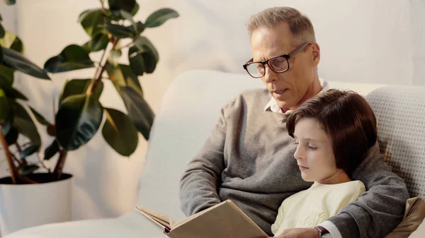 Middle aged grandfather in glasses reading book to grandchild at home — стоковое фото