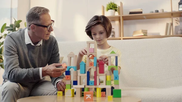 Abuelo de mediana edad en gafas mirando nieto jugando bloques de construcción - foto de stock