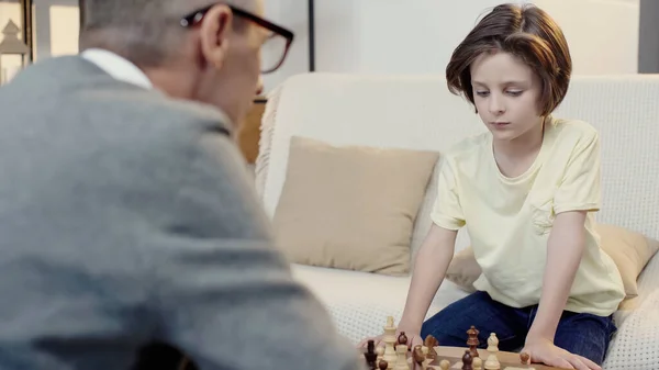 Boy playing chess with blurred grandfather in living room — стоковое фото