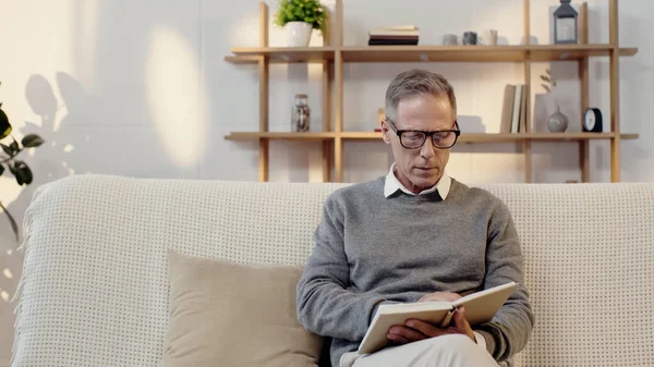 Чоловік середнього віку в окулярах читає книгу у вітальні — стокове фото