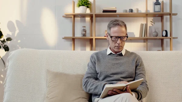 Middle aged man in glasses reading book in living room — стоковое фото