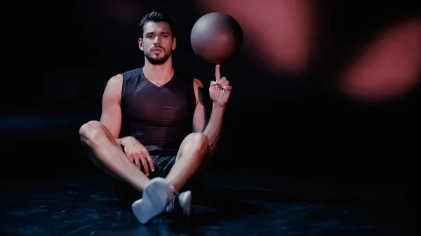 Sportsman spinning basketball on finger while sitting on black — Stock Photo