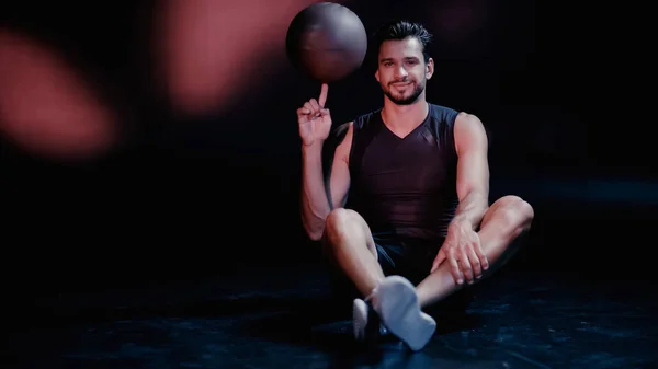 Happy sportsman spinning basketball on finger while sitting on black — Stock Photo