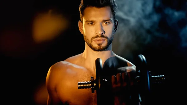 Shirtless and muscular sportsman training with dumbbell near smoke on black — Stock Photo