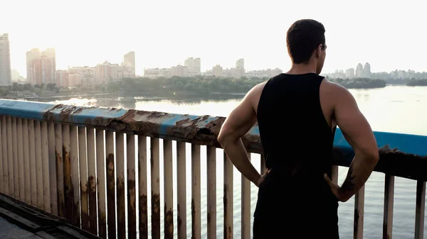 Vue arrière du sportif mixte debout avec les mains sur les hanches sur le pont au-dessus de la rivière — Photo de stock