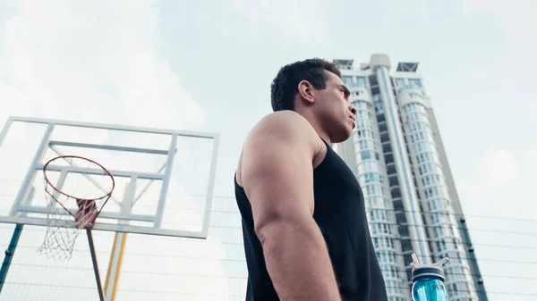 Low angle view of sportive mixed race man with sports bottle near basketball ring outdoors — Stock Photo