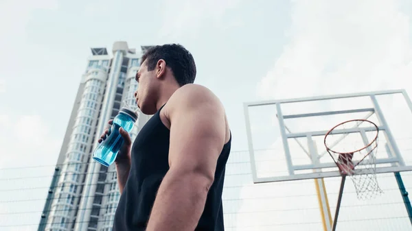 Vista de ángulo bajo del hombre atlético bi-racial sosteniendo botella deportiva cerca del anillo de baloncesto al aire libre - foto de stock