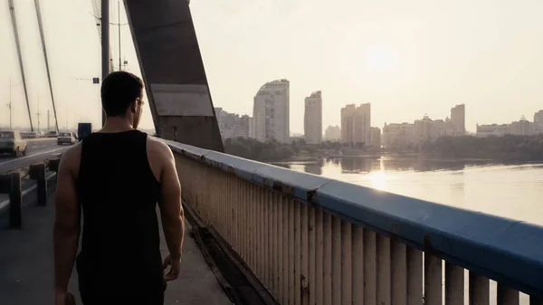 Назад вид спортивной смешанной расы человека, идущего по городскому мосту через реку — стоковое фото