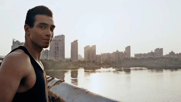 Sport bi-razziale uomo guardando la fotocamera sul ponte della città sul fiume — Foto stock