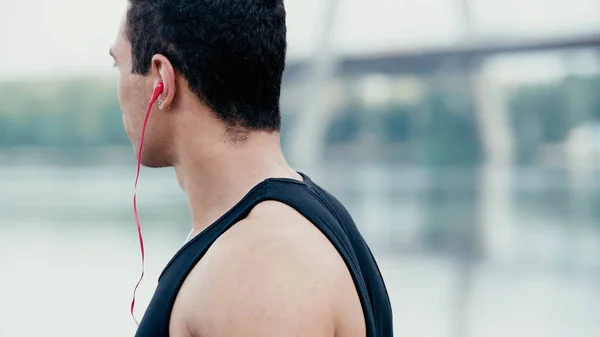Jovem e esportivo bi-racial homem em fones de ouvido ouvindo música ao ar livre perto do rio turvo — Fotografia de Stock
