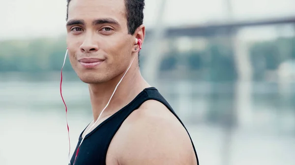 Athletic bi-racial man in earphones smiling at camera near blurred river — Stock Photo