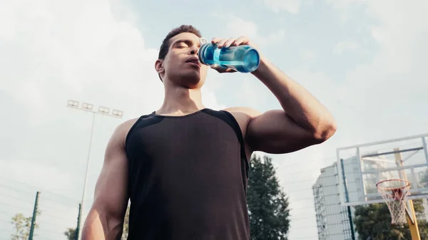 Низкий угол обзора двурасового спортсмена питьевой воды из спортивной бутылки на открытом воздухе — стоковое фото