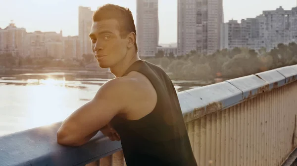 Atlético bi-racial hombre de pie en el puente de la ciudad sobre el río y mirando a la cámara - foto de stock