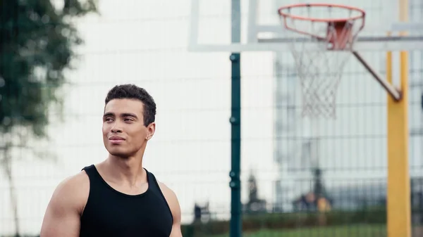 Homme bi-racial athlétique regardant loin près de l'anneau flou de basket-ball à l'extérieur — Photo de stock