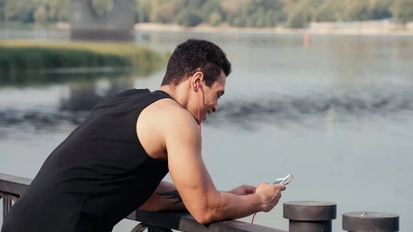 Hombre bi-racial feliz y deportivo en auriculares usando teléfono inteligente en el puente sobre el río - foto de stock