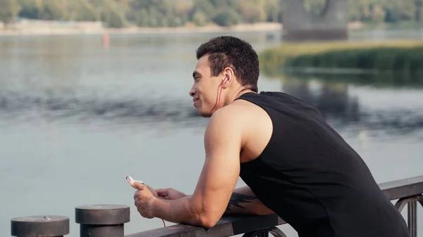 Sonriente hombre bi-racial con teléfono inteligente escuchar música en el puente sobre el río - foto de stock