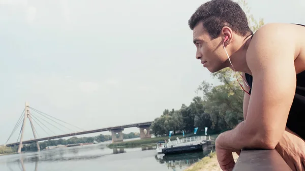 Вид сбоку спортивного бирасового человека, слушающего музыку, глядя на реку — стоковое фото
