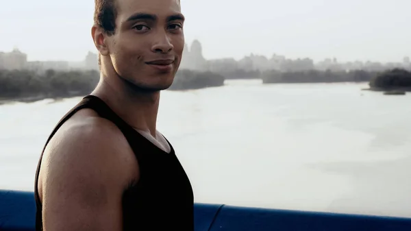 Happy bi-racial sportsman looking at camera while standing on bridge over river — Stock Photo