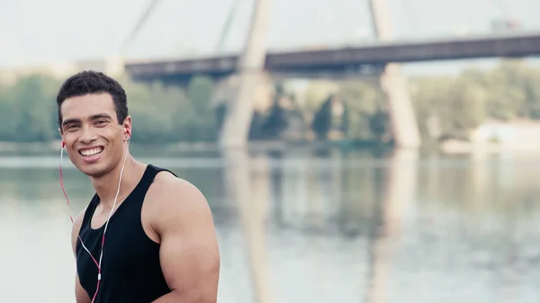 Позитивний змішаний гоночний спортсмен в навушниках, що дивляться на камеру біля річки та розмитого мосту — стокове фото