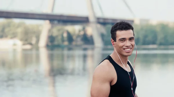 Bi-racial sportsman looking at camera near bridge over river on blurred background — Stock Photo