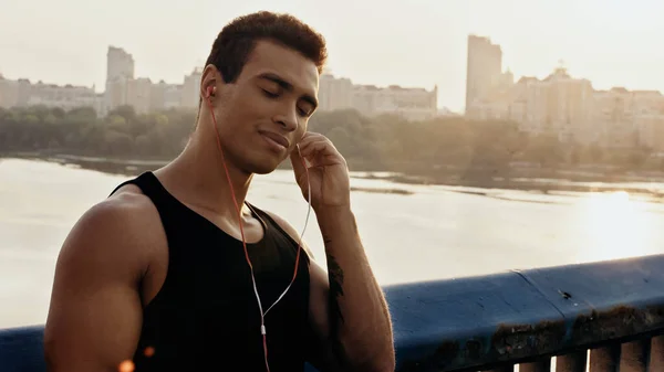 Sorrindo homem de raça mista com olhos fechados ouvindo música em fones de ouvido na ponte sobre o rio — Fotografia de Stock