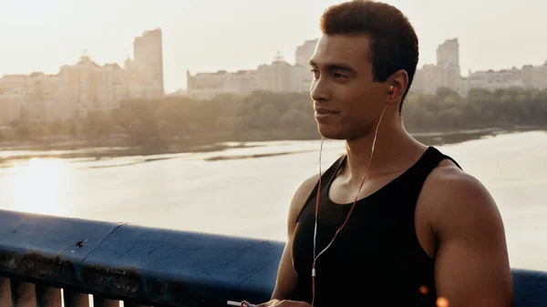 Smiling bi-racial sportsman in earphones listening music on city bridge over river — Stock Photo