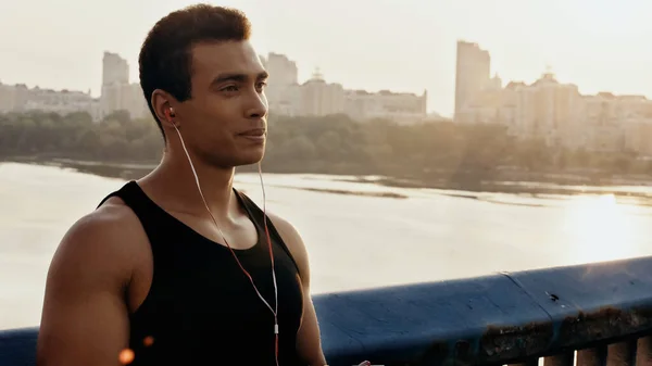 Joven y atlético hombre de raza mixta escuchando música en el puente sobre el río - foto de stock