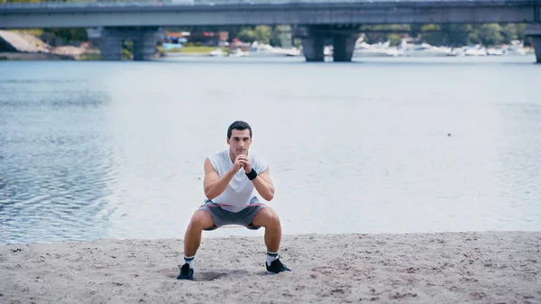 Joven deportista atlético haciendo ejercicio en cuclillas en la orilla del río - foto de stock