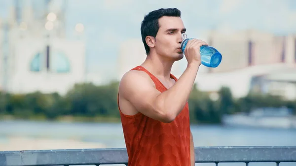 Junger Sportler steht auf Brücke und trinkt Wasser aus Sportflasche — Stockfoto