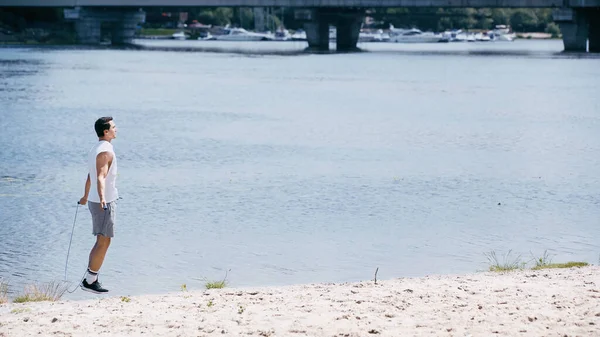 Seitenansicht eines jungen Sportlers, der mit Springseil auf Sand in Flussnähe springt — Stockfoto