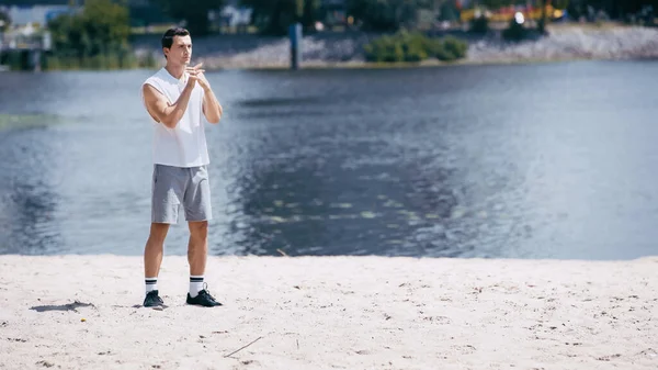 Joven deportista en camiseta sin mangas y pantalones cortos calentando los dedos cerca del río - foto de stock