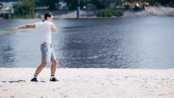 Young sportsman in tank top and shorts doing body rotation near river — Stock Photo