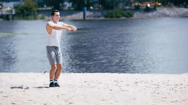 Young sportsman in tank top and shorts warming up hands near river — Stock Photo