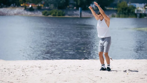 Joven deportista en camiseta sin mangas y pantalones cortos calentándose con las manos extendidas cerca del río - foto de stock