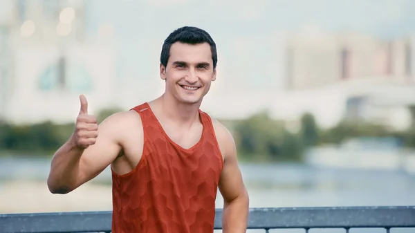 Atleta atlético feliz sorrindo e mostrando o polegar para cima enquanto está em pé na ponte — Fotografia de Stock