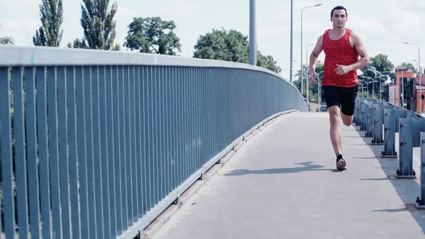 Young sportsman in tank top and shorts running on bridge in summer — Stock Photo