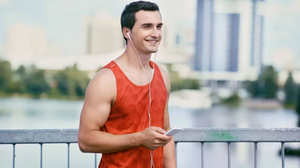 Happy sportsman holding smartphone and listening music in earphones — Stock Photo