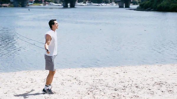 Side view of young man jumping with skipping rope on sand near river — Stock Photo