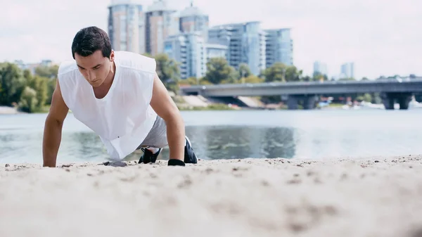 Young sportsman doing push ups on sand near river — Stock Photo