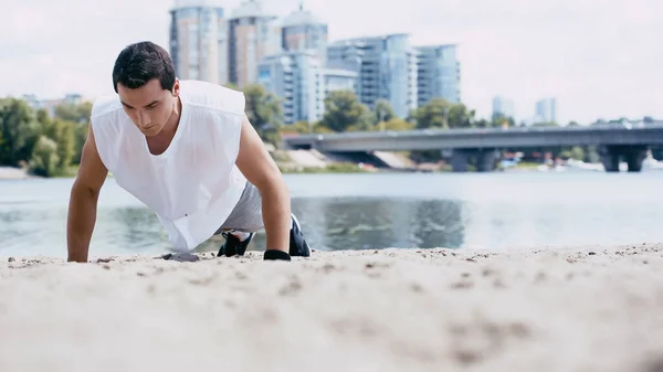 Joven deportista haciendo flexiones en la arena cerca del río - foto de stock