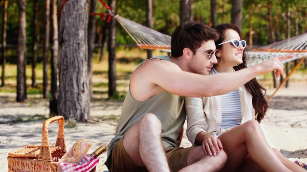 Hombre en gafas de sol señalando con el dedo cerca de novia sonriente y cesta de picnic en la playa - foto de stock