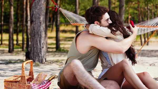 Mann hält Schmuckschatulle und umarmt Freundin in der Nähe von Picknickkorb am Strand — Stockfoto