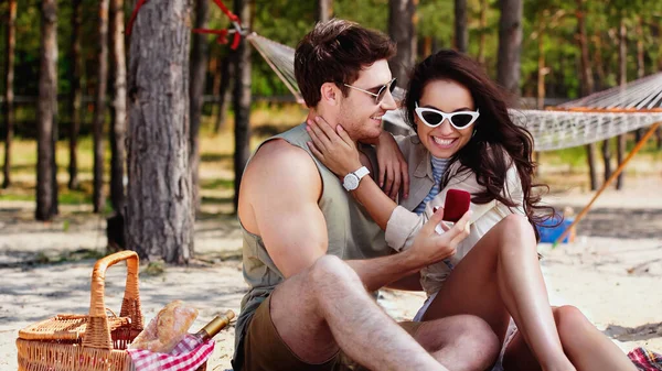 Smiling woman in sunglasses touching boyfriend with jewelry box during picnic on beach — Stock Photo