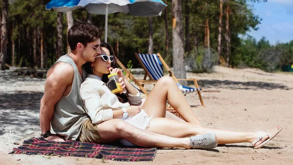 Woman in sunglasses drinking orange juice near boyfriend on beach — Stock Photo