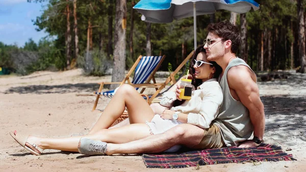 Улыбающаяся женщина в солнечных очках держит апельсиновый сок рядом с парнем на одеяле на пляже — стоковое фото